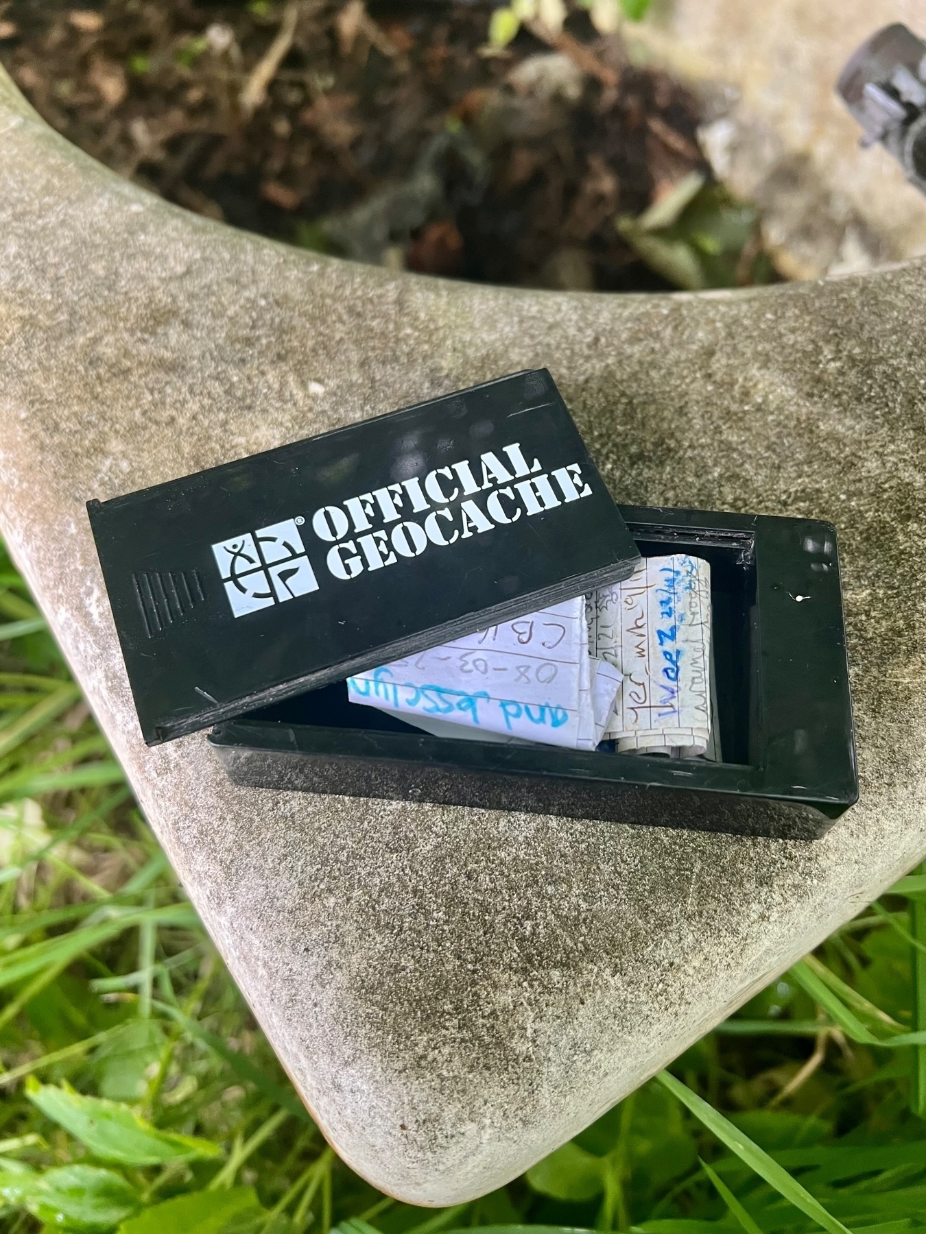 A small black container labeled Official Geocache is open, revealing a stack of papers inside, placed on a stone surface surrounded by grass.