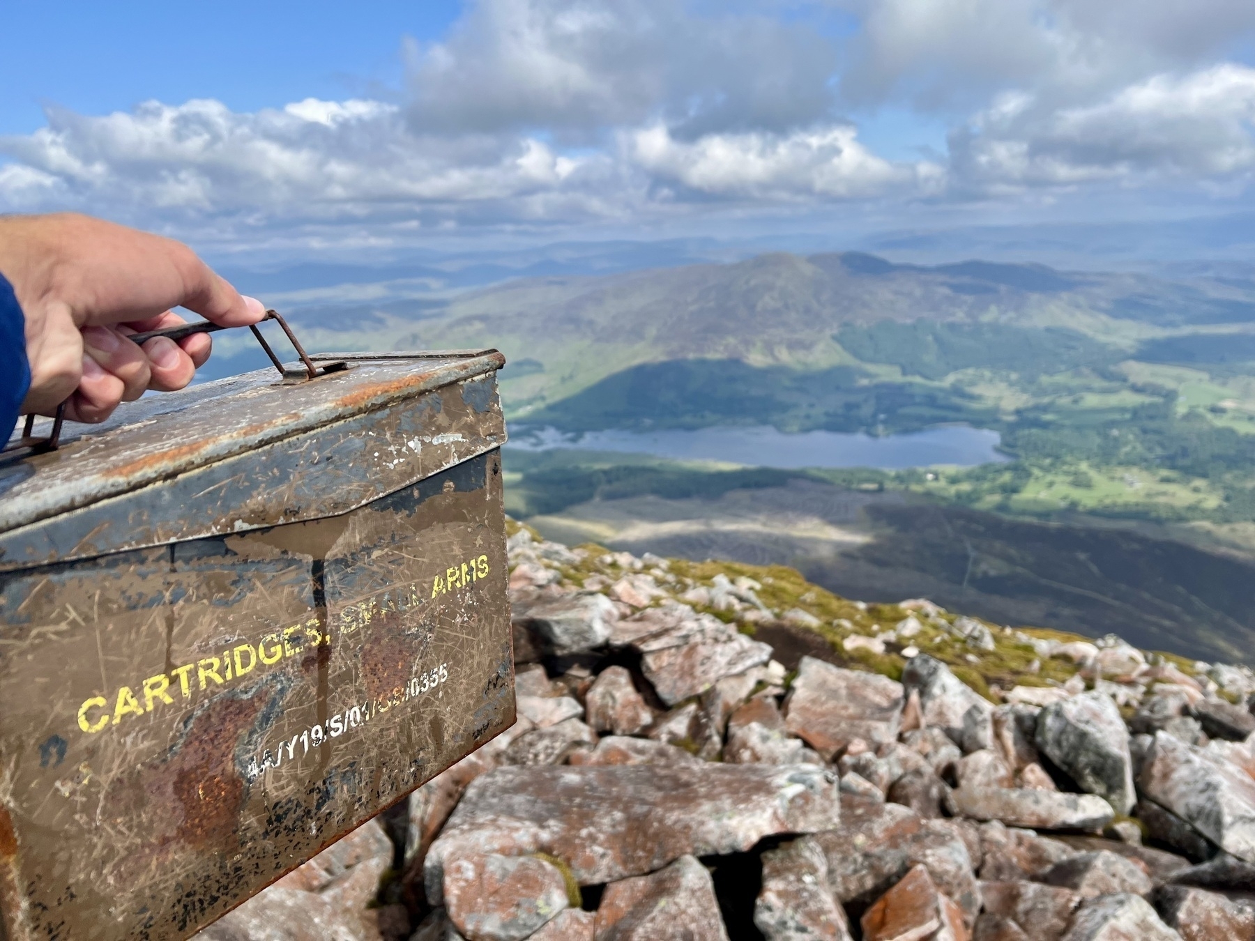 A person is holding a rusty ammunition box labeled CARTRIDGES SMALL ARMS atop a rocky mountain with a scenic view of a valley, lake, and mountains in the background.
